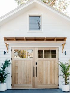 a white house with two wooden garage doors and plants in pots on the front porch