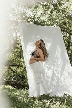 a pregnant woman standing in front of a white backdrop with trees and grass around her