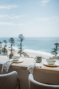 two cups and saucers are sitting on a window sill overlooking the beach with pine trees