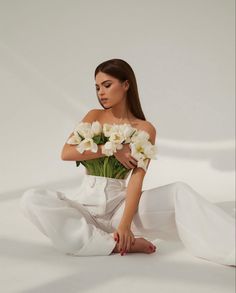 a woman sitting on the ground with flowers in her hand and wearing a white dress