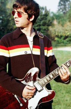 a young man holding an electric guitar in front of some trees and grass with sunglasses on