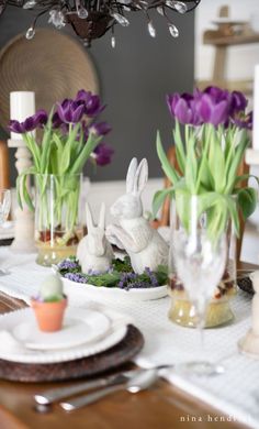 the table is set for easter dinner with purple tulips and white bunny figurines