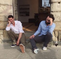 two men sitting on the ground next to each other with wine glasses in front of them