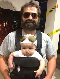 a man is holding a baby in a black carrier and wearing sunglasses on his head