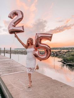 a woman holding two large balloons in the shape of numbers on a dock at sunset