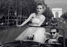 a woman in a white dress is riding in a convertible car with a man standing next to her