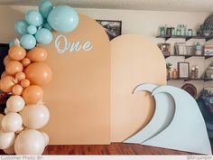 balloons are hanging on the wall in front of a surfboard and balloon arch that says one