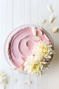 a pink frosted cake with white flowers on the side and petals scattered around it