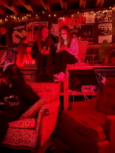 two women sitting on couches in a room with red light from the ceiling and posters above them