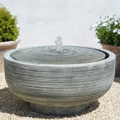 a water fountain in the middle of some potted plants and graveled area next to a building