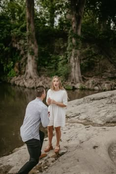 a man kneeling down next to a woman on top of a rock near the water