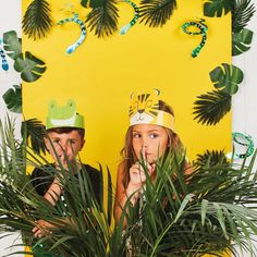 two people wearing party hats standing in front of a yellow backdrop with palm trees and foliage