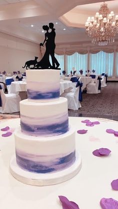 a white and blue wedding cake with purple flowers on the table in front of it