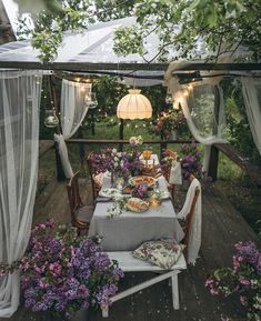 an outdoor dining area with table and chairs covered in curtains, flowers and lights hanging from the ceiling