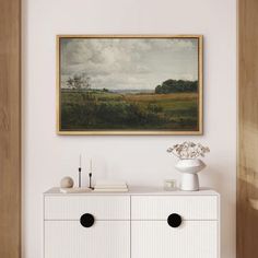 a painting hanging on the wall above a white dresser with two drawers and a vase filled with flowers