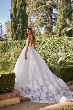 a woman in a white wedding dress standing on some steps near bushes and trees with her back turned to the camera