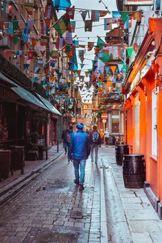 people walking down an alley way with colorful flags hanging from the buildings above them,