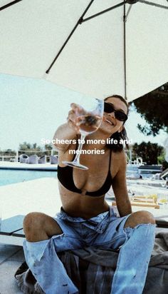 a woman sitting on the ground drinking from a wine glass with an umbrella in the background