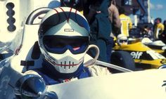 a man sitting in a race car wearing a helmet and goggles with other people around him