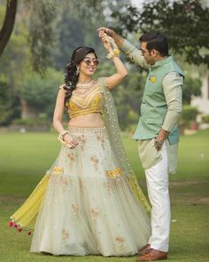 a man standing next to a woman in a white and yellow lehenga on top of a lush green field