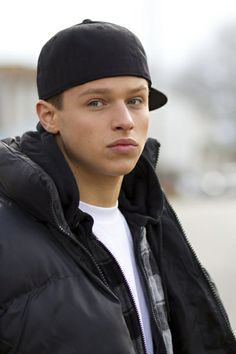 a young man in a black jacket and hat looking at the camera with a serious look on his face