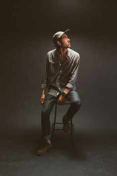 a man sitting on top of a chair in front of a black background wearing a hat