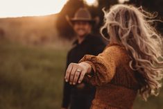 a man and woman holding hands while walking through a field with the sun behind them