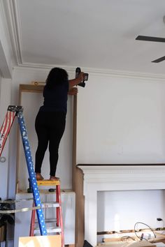 a woman standing on top of a ladder in front of a fire place and ceiling fan