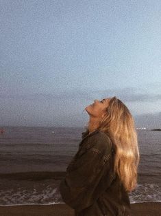 a woman standing on top of a beach next to the ocean looking up at a kite flying in the sky