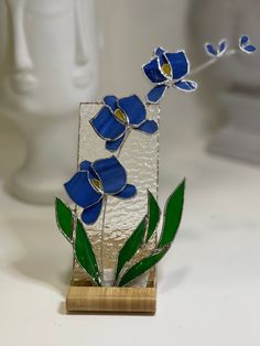 a glass vase with blue flowers and green leaves in it sitting on a wooden stand