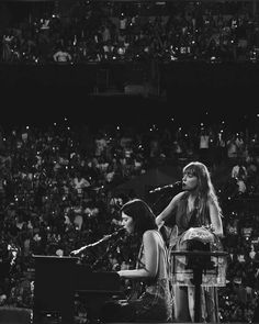 two women are sitting at a piano in front of an audience and one is singing into a microphone