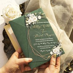 two hands holding a green and gold wedding card with white flowers on it next to a bouquet of roses