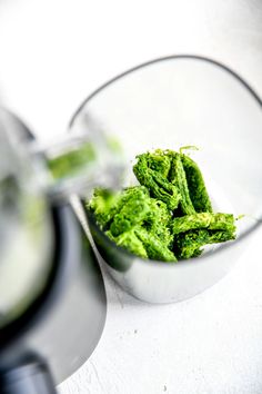 a blender filled with green vegetables on top of a white counter next to a metal bowl