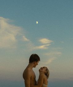 a man and woman standing on the beach looking at each other under a full moon