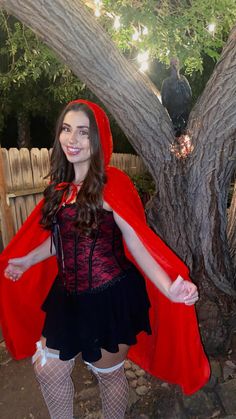 a woman in a red cape and black dress standing next to a tree with her arms outstretched