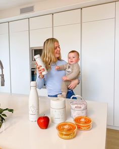 a woman holding a baby in her arms while standing next to some food on a kitchen counter