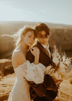 a man and woman standing next to each other on a dirt field with mountains in the background