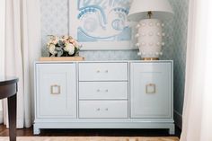 a blue dresser with flowers on top and a painting above it in a room that has white curtains