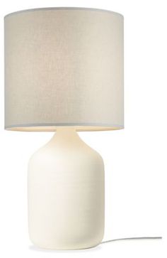 a white table lamp with a beige shade on the base and a light bulb plugged in