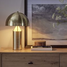 a lamp on top of a wooden dresser next to a painting and a vase with flowers