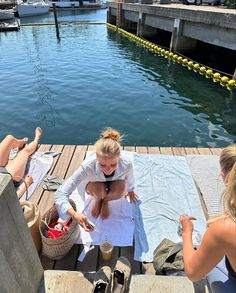 three people sitting on a dock next to the water and one woman is tying her tie