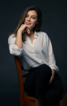 a woman sitting on top of a wooden chair in front of a dark background with her hand under her chin