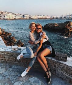 two women are sitting on a stone wall by the water and taking a selfie