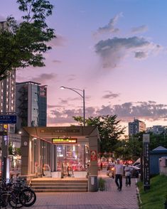 people are walking down the sidewalk in front of a building at dusk with tall buildings behind them