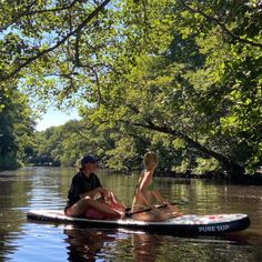 two people sitting on a paddle board in the middle of a river with trees around them