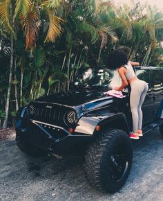 a woman leaning on the hood of a black jeep parked in front of palm trees