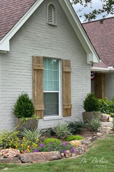 a white house with wooden shutters and flowers in the front yard on a sunny day