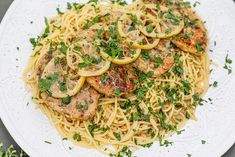 a white plate topped with pasta covered in chicken and lemon slices next to parsley