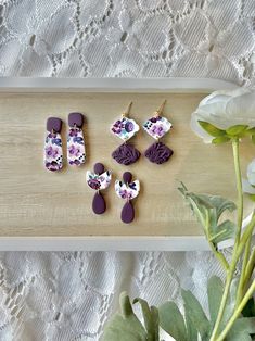 purple and white flowers are sitting on a wooden tray next to small earrings that have been made from polymer