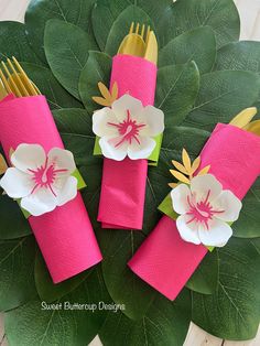 three pink napkins with white flowers on them and gold forks in the middle are surrounded by green leaves